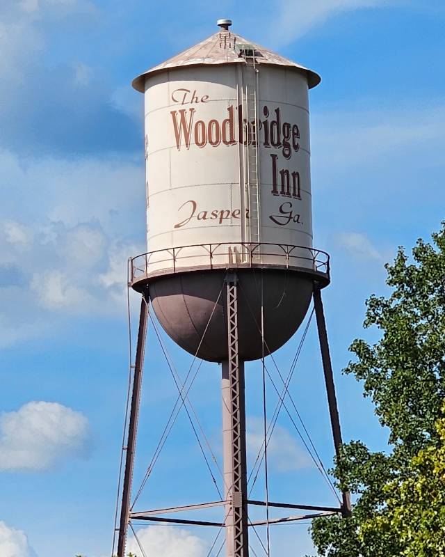 A photo taken from downtown Jasper Georgia of The Woodbridge Inn water tower. The entire photo is of the top of the water tower. The water town is dark brown, with it's body painted tan. It is an old style water town which features a cylinder body, and a cone top. It rests on 4 large legs with a pipe in the middle. The text on the side of the water tower reads "The Woodbridge Inn Jasper GA." The background is of a partly cloudy blue sky and the bottom right features the foliage of a nearby tree. Jasper GA is the hometown of Pinwheel Web Development